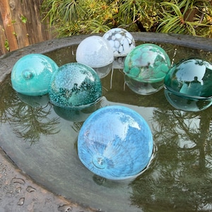 May include: A collection of seven spherical glass floats in various shades of blue, green, and white, resting in a dark circular basin filled with water. The glass orbs feature diverse textures, including solid colors, speckled patterns, and translucent finishes.