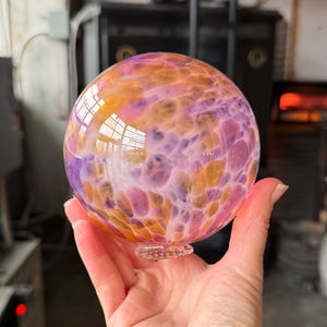 May include: A colorful, spherical glass art piece. The orb features a blend of orange, purple, and pink hues. The glass is transparent, allowing light to pass through and reflect off the surface. The bottom has a small, decorative base.
