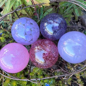 May include: A collection of five decorative glass spheres in a wire basket. The spheres are various shades of purple and pink, with some featuring textured or speckled designs. The spheres are resting on a bed of green foliage.