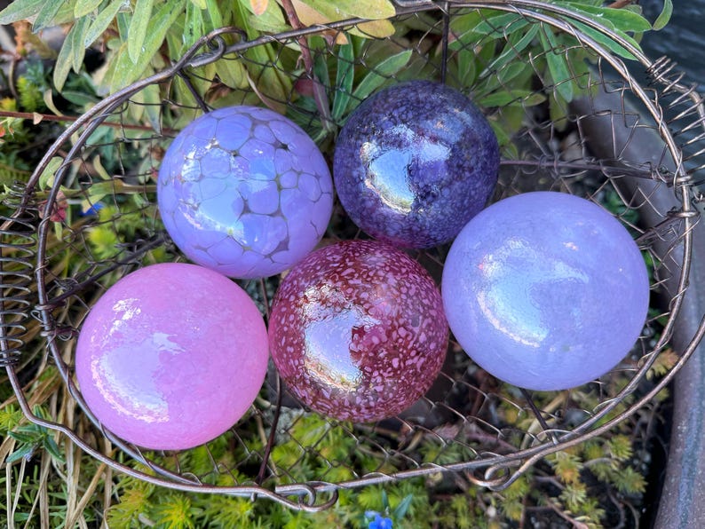 May include: A collection of five decorative glass spheres in a wire basket. The spheres are various shades of purple and pink, with some featuring textured or speckled designs. The spheres are resting on a bed of green foliage.