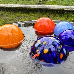 May include: Four glass orbs, two orange, one blue, and one blue with yellow and orange accents, float in a shallow pool of water.