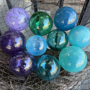 May include: A collection of ten spherical glass orbs in various shades of purple, blue, and green. The spheres feature smooth, reflective surfaces with some displaying internal crackle patterns or deep, saturated hues, arranged together in a wire basket.