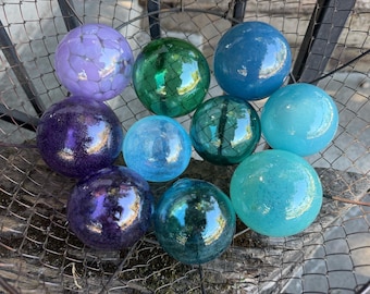 Puede incluir: Una colección de diez esferas de vidrio en varios tonos de morado, azul y verde. Las esferas presentan superficies lisas y reflectantes, algunas con patrones internos de grietas o tonos saturados profundos, dispuestas juntas en una cesta de alambre.