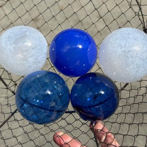 May include: Five spherical glass orbs arranged in a wire mesh basket. Two orbs feature a translucent white mottled pattern, one is a solid vibrant blue, and two are a deep, dark blue with a smooth, reflective surface finish and clear glass texture.