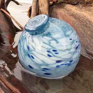 May include: A round, blue and white glass vase with a decorative stopper. The vase has a swirl pattern with dark blue spots. The stopper has a spiral design. The vase is sitting on a piece of driftwood.