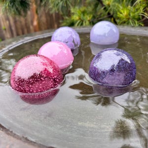 May include: A collection of colorful, spherical bath bombs floating in water. The bath bombs are various shades of pink, purple, and lavender, with a speckled texture. The water is in a dark, circular basin, with greenery in the background.