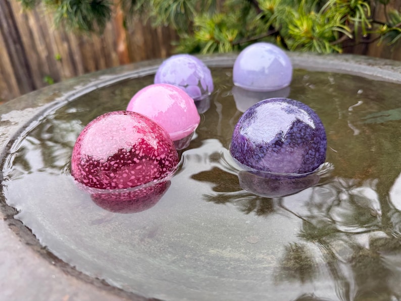 May include: A collection of colorful, spherical bath bombs floating in water. The bath bombs are various shades of pink, purple, and lavender, with a speckled texture. The water is in a dark, circular basin, with greenery in the background.