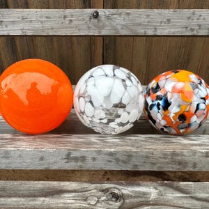 May include: Three decorative glass spheres in a row. The first is solid orange, the second is white with black and gray patterns, and the third is a mix of orange, black, and blue. They rest on a weathered wooden surface.