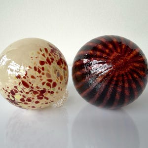 May include: Two decorative glass spheres. One is beige with red and yellow speckles. The other is dark red with a spiral pattern of dark brown lines. Both spheres are round and appear to be handmade.