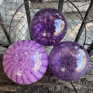 May include: Three decorative glass spheres in shades of purple, resting in a metal wire basket. The spheres have textured surfaces and varying patterns, with one displaying a swirl design. The basket is made of dark metal.