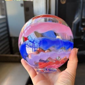 May include: A spherical glass ornament featuring horizontal wavy bands of color in pink, blue, purple, and red. The clear glass orb has a smooth, polished surface with a small circular base at the bottom, showcasing layered translucent color patterns.