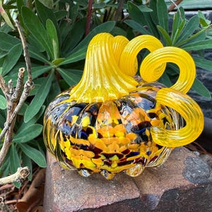 May include: A decorative glass pumpkin with a yellow stem and curly tendrils. The body of the pumpkin is a mix of yellow, orange, and black, with hints of blue. The pumpkin is sitting on a stone surface, surrounded by green foliage.