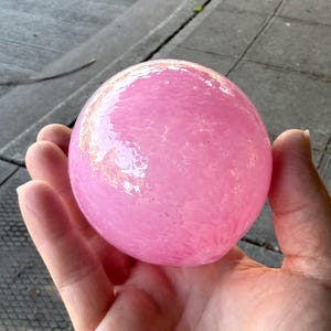 May include: A pink, spherical object with a textured surface. The object is held in an open hand, with the background showing a gray surface. The sphere appears to be made of glass or a similar material.