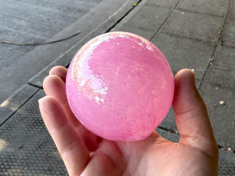 May include: A pink, spherical object with a textured surface. The object is held in an open hand, with the background showing a gray surface. The sphere appears to be made of glass or a similar material.
