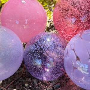 May include: A collection of decorative glass spheres in various shades of pink and purple. The spheres have unique textures and patterns, with some appearing speckled and others smooth. These colorful orbs are ideal for garden decor.