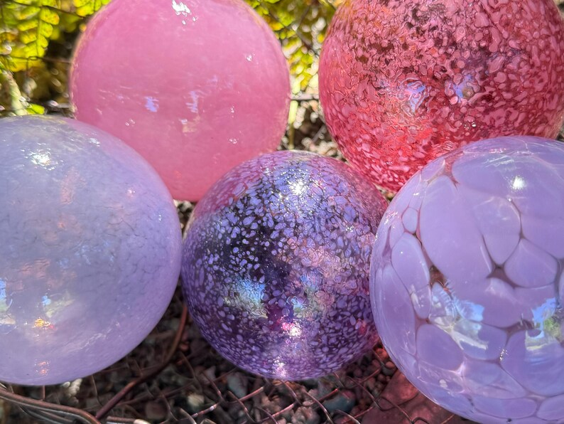 May include: A collection of decorative glass spheres in various shades of pink and purple. The spheres have unique textures and patterns, with some appearing speckled and others smooth. These colorful orbs are ideal for garden decor.