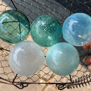 May include: Five spherical glass floats in shades of teal, light blue, and white, resting inside a dark brown wire basket. The glass spheres feature varied textures, including smooth, crackled, and mottled surfaces, arranged in a cluster.