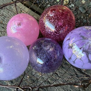 May include: Five colorful glass spheres in shades of pink, purple, and burgundy rest in a metal basket. The spheres have unique patterns and textures, with some appearing speckled and others with floral designs.