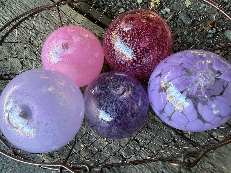 May include: Five colorful glass spheres in shades of pink, purple, and burgundy rest in a metal basket. The spheres have unique patterns and textures, with some appearing speckled and others with floral designs.