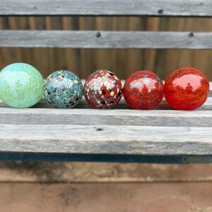 May include: Five colorful glass spheres in shades of green, blue, red, and orange are arranged in a row. The spheres have unique patterns and textures, with some appearing speckled and others smooth. They rest on a weathered wooden surface.