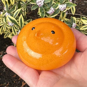 May include: A round, orange glass paperweight with two small black dots and a curved indentation, resembling a smiling face. The paperweight is held in a hand, with a blurred background of green and yellow foliage.