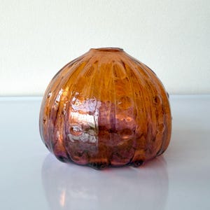 May include: A decorative glass vase with a bulbous shape and a small opening at the top. The vase is a warm amber color with a textured surface and small, round indentations. The base has a hint of purple.