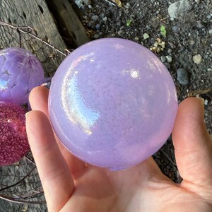 May include: A hand holds a large, light purple glass sphere with a speckled texture. Other colorful glass spheres, including pink and purple, are visible in a wire basket. The spheres have a decorative, iridescent quality.