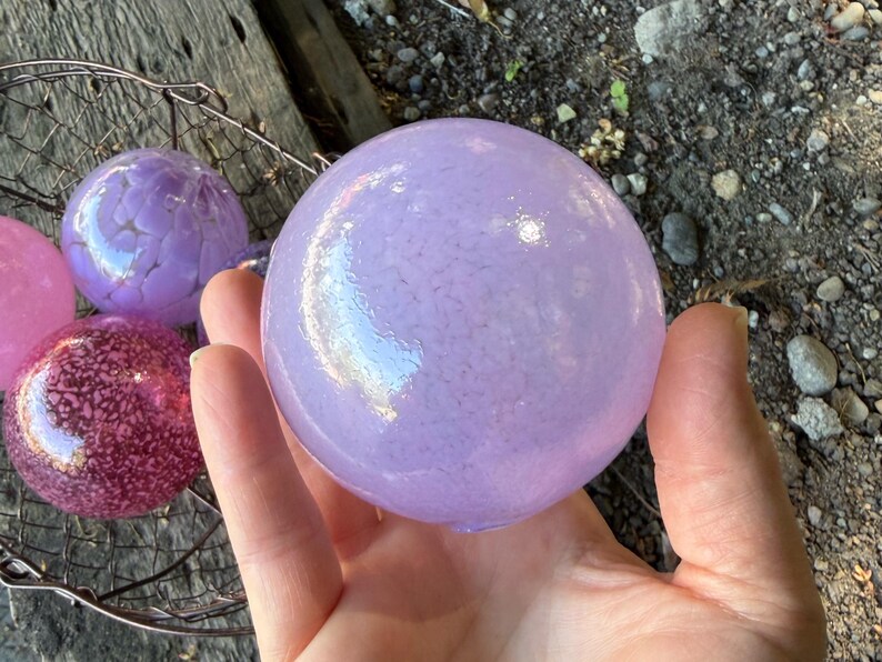 May include: A hand holds a large, light purple glass sphere with a speckled texture. Other colorful glass spheres, including pink and purple, are visible in a wire basket. The spheres have a decorative, iridescent quality.
