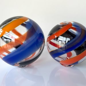 May include: Two spherical glass paperweights featuring clear glass bodies with swirling bands of orange, blue, and black colors. The spheres are smooth and reflective, showcasing a layered design where the colored ribbons wrap around the interior.