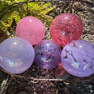 May include: A collection of five decorative glass spheres in a rustic metal basket. The spheres vary in shades of pink, purple, and lavender, with speckled and patterned designs. The spheres are a decorative garden or home accent.
