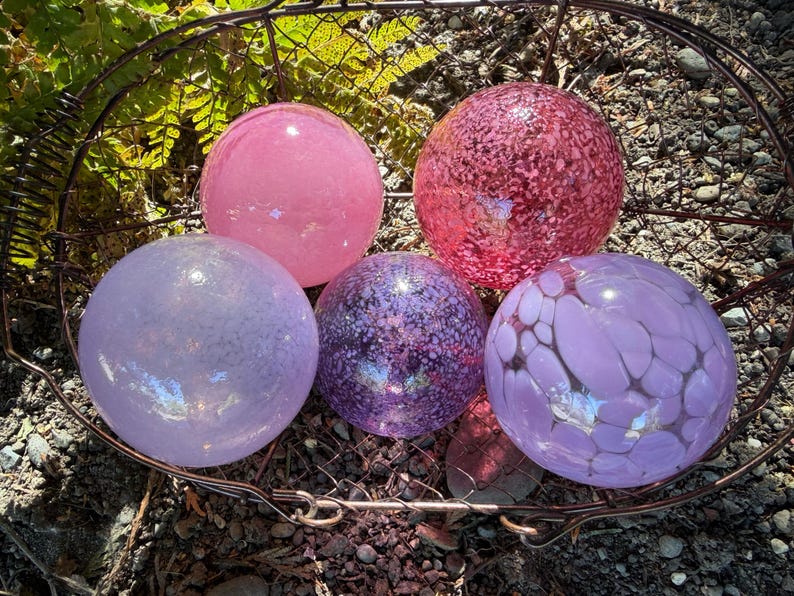 May include: A collection of five decorative glass spheres in a rustic metal basket. The spheres vary in shades of pink, purple, and lavender, with speckled and patterned designs. The spheres are a decorative garden or home accent.