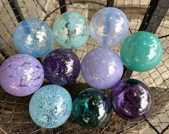May include: A collection of ten glass ornaments in various shades of blue, purple, and green. The ornaments have a textured, iridescent finish and are arranged in a circular pattern.