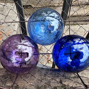 May include: Three spherical glass orbs with a crackled texture resting inside a dark metal wire basket. One orb is a deep purple, while the other two are shades of blue, ranging from a lighter sky blue to a vibrant, saturated royal blue color.