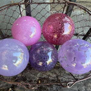 May include: Five decorative glass spheres in shades of pink, purple, and lavender, displayed in a rustic wire basket. The spheres have varying textures, including smooth and patterned surfaces, and reflect light.