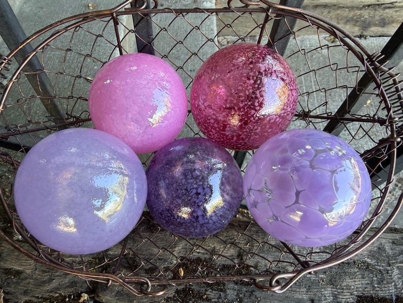 May include: Five decorative glass spheres in shades of pink, purple, and lavender, displayed in a rustic wire basket. The spheres have varying textures, including smooth and patterned surfaces, and reflect light.