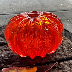 May include: A vibrant, round, orange glass pumpkin with vertical ridges. The decorative piece has a small opening at the top and sits on a weathered wooden surface. The pumpkin's color is a bright, translucent orange.