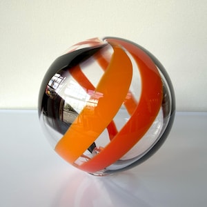 May include: A clear glass sphere with orange, red, and black swirling stripes. The decorative orb reflects light and has a glossy finish. The sphere is a decorative art object.
