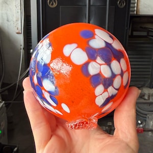 May include: A spherical glass ornament featuring a vibrant orange base color with scattered, rounded patches of opaque white and deep blue glass. The surface is smooth and glossy, with a small, textured glass nub at the bottom of the sphere.