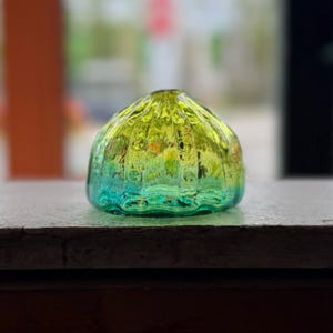May include: A small, decorative glass object with a rounded shape. The glass transitions from a vibrant yellow-green at the top to a deep teal at the base. The surface has a textured, bubbly appearance, and it sits on a neutral surface.