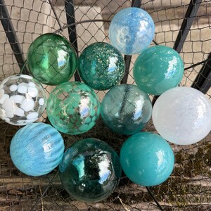 May include: A collection of eleven decorative glass spheres in various shades of blue, green, and white. The spheres feature diverse textures, including clear glass with white spots, solid opaque finishes, translucent green, and glittery surfaces.