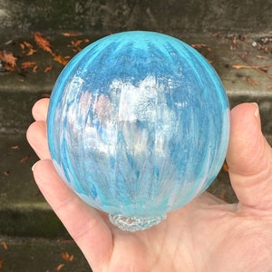 May include: A round, glass ornament with a light blue and white color scheme. The ornament has a textured, painted design and a small, decorative glass base. The ornament is held in a hand.