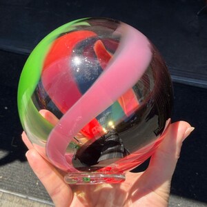 May include: A spherical glass paperweight featuring swirling ribbons of translucent lime green, vibrant red, and soft pastel pink encased within a clear glass orb. The object has a smooth, polished surface and a small, flat circular base for stability.