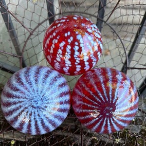 May include: Three decorative glass spheres in a wire basket. One sphere is red with white spots, and two are red and blue with striped patterns. The spheres are a vibrant addition to any home decor.