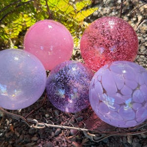 May include: Five decorative glass spheres in shades of pink and purple rest in a wire basket. The spheres vary in texture, with some appearing smooth and others speckled or patterned. The arrangement is set against a backdrop of green foliage.