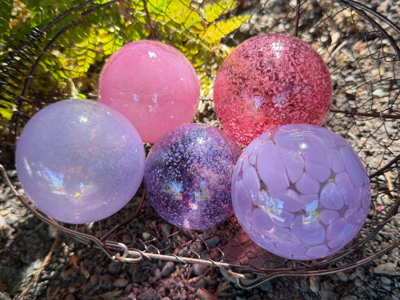 May include: Five decorative glass spheres in shades of pink and purple rest in a wire basket. The spheres vary in texture, with some appearing smooth and others speckled or patterned. The arrangement is set against a backdrop of green foliage.