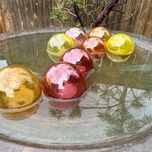 May include: A bird bath filled with water and floating glass orbs in shades of yellow, orange, and red.