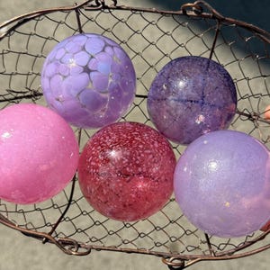 May include: Five colorful glass spheres in shades of pink, purple, and red are displayed in a wire basket. The spheres have various textures, including speckled and smooth surfaces. The basket is made of dark metal.