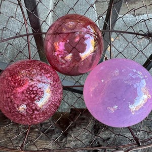 May include: Three glass ornaments in a wire basket. The ornaments are pink, purple, and a darker shade of pink. The ornaments have a textured, crackled finish.