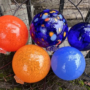 May include: Five glass ornaments in various colors and patterns. The ornaments are blue, orange, and light blue. Some of the ornaments have a speckled pattern, while others have a smooth surface.