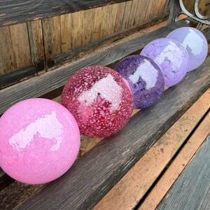 May include: Six decorative glass spheres in shades of pink, red, purple, and lavender are arranged on a wooden bench. The spheres have textured surfaces and reflect light, creating a visually appealing display. The spheres vary in size and color.